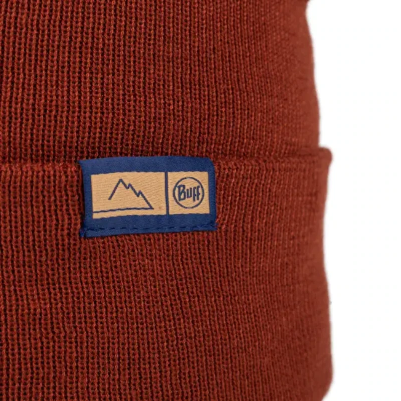 Un primer plano del Gorro Buff Knitted Beanie Lilon Long revela su color rojo y una etiqueta rectangular azul con un gráfico de montaña con "Buff" sobre un fondo beige, que incorpora un acogedor estilo invernal.