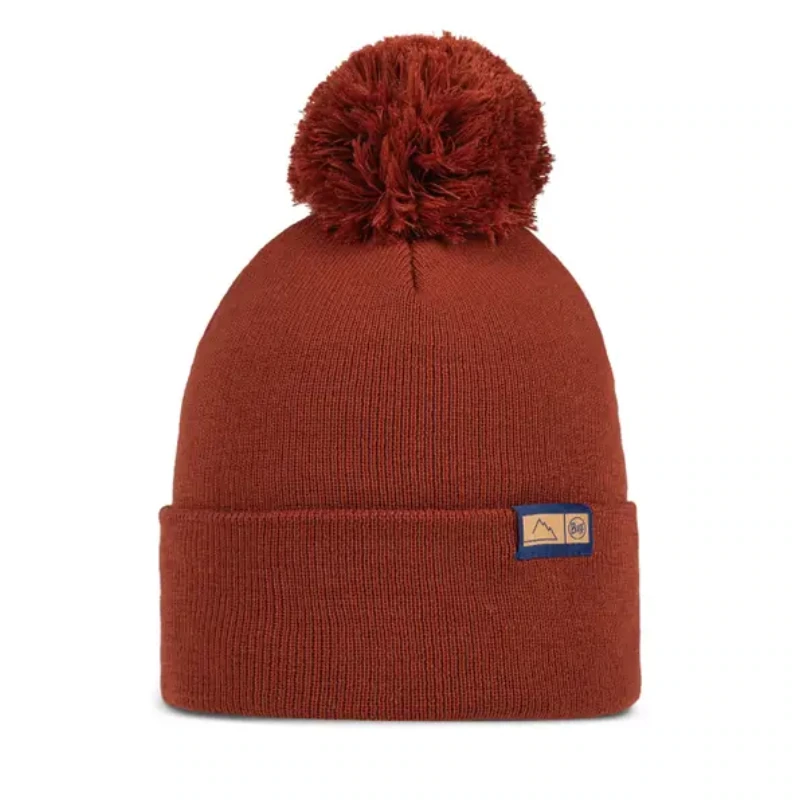 Un gorro de punto Gorro Buff Lilon Long de color óxido con un pompón grande presenta un pequeño parche rectangular con diseños de montañas y copos de nieve en el frente.
