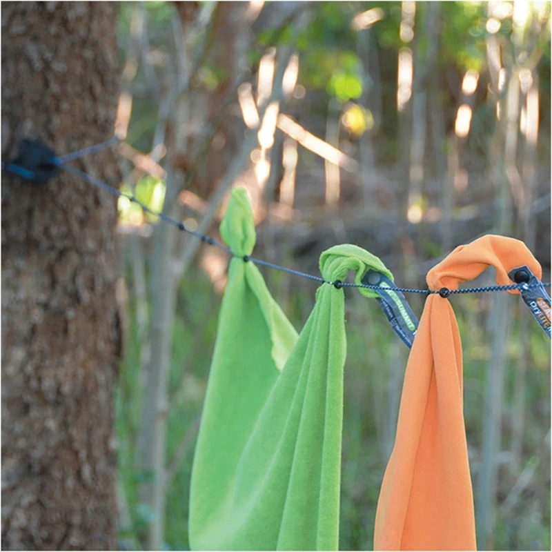 Una toalla verde y naranja cuelga de un tendedero de Sea to Summit, con mosquetones para accesorios, entre los árboles de un bosque. Troncos de árboles y follaje borrosos forman el fondo.