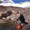 Una persona vestida de negro se agacha sobre un terreno rocoso, usando un balde plegable rojo Cubeta Plegable Sea To Summit y una olla para recoger agua de una pequeña piscina. Cerca de allí, su Silla Inflable Sea to Summit Air Chair espera mientras las montañas nevadas se alzan contra el cielo azul claro.