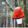 Una bolsa naranja con símbolos de baños cuelga de la rama de un árbol en un bosque, similar al Tendedero Portátil Sea to Summit The Clothesline. Una tela o papel blanco ondea sobre la bolsa entre un follaje verde exuberante y troncos robustos.