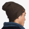 La persona lleva un gorro Buff Niños Knitted y Polar Lyne marrón con un pequeño parche de cuero, combinado con una chaqueta de polar azul. Vistos de lado sobre un fondo blanco liso, destilan un estilo informal.