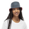Una persona con el pelo largo trenzado sonríe mientras lleva un sombrero de pescador Cinta Buff Fastwick Barriers Graphite de color azul oscuro y una camiseta blanca con la marca Cinta. El fondo blanco liso resalta su estilo sin esfuerzo.