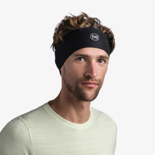 Un hombre con cabello castaño corto y rizado y barba lleva una cinta para el pelo negra con el logotipo Cinta Buff CoolNet UV Wide Solid y una camiseta verde claro de manga larga. Está de pie sobre un fondo blanco liso.
