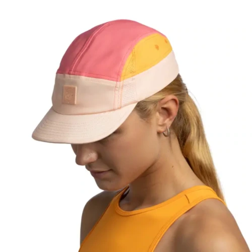Una persona que lleva una gorra Jockey Buff Speed Pink Deri con secciones rosas, naranjas y beige mira hacia abajo. Tiene el pelo largo y rubio y lleva una camiseta sin mangas naranja sobre un fondo blanco liso.