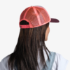 Se ve a una persona desde atrás vistiendo una gorra de béisbol Jockey Buff Trucker Green Campast con una correa ajustable, cabello largo y oscuro cayendo en cascada y vestida con una camiseta blanca combinada con un mono azul.