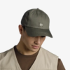 La persona lleva una gorra de béisbol Jockey Buff Zire verde oscuro con el logotipo "Buff". Tiene un chaleco beige sobre una camisa marrón y mira hacia abajo sobre un fondo blanco.