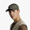 Un joven viste una gorra de béisbol Jockey Buff Baseball Zire verde oscuro, con un logotipo, con un chaleco beige sobre una camisa marrón, mirando con confianza a la cámara contra un fondo gris liso.
