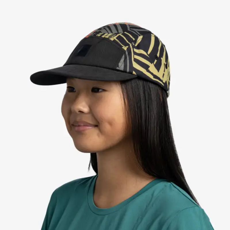 Una persona con cabello largo y oscuro lleva una camiseta verde azulado y una gorra Jockey Buff 5 Panel Go Pow Running Multi con patrones abstractos dorados y negros. Sonríe, mira ligeramente hacia la derecha y exuda un aire de aventura sobre un fondo blanco liso.