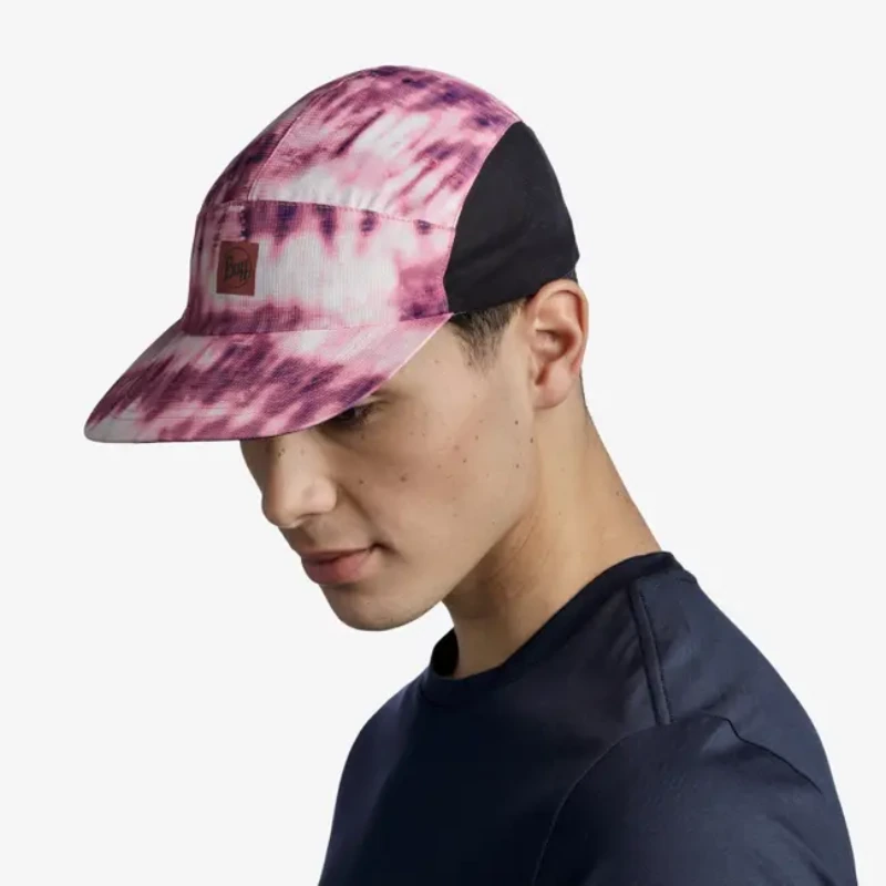 Una persona lleva una gorra Jockey Buff Speed Pink Deri, teñida en rosa y blanco con secciones laterales negras y una etiqueta marrón, y la combina con una camisa oscura mientras mira hacia abajo pensativamente sobre un fondo blanco liso.