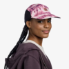Con la gorra Jockey Buff Speed Pink Deri, una persona con cabello oscuro trenzado y una sudadera con capucha oscura sonríe sutilmente, mirando de reojo contra un fondo sencillo, emanando vibraciones geniales.