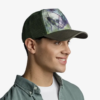 Una persona sonriente con una gorra de béisbol con un diseño abstracto y una camisa verde abotonada mira hacia la derecha. La gorra Jockey Buff Trucker Wadi Fawn (copia) se destaca sobre el fondo blanco liso.