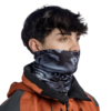 Con una chaqueta naranja y negra, una persona de pelo corto y oscuro mira de reojo. Una bandana Buff ThermoNet Skatick Graphite le cubre la mitad inferior del rostro, mostrando los logotipos de la marca sobre un fondo blanco puro.