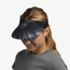 Una persona con cabello largo lleva una elegante visera negra y gris con un patrón geométrico. Está vestida con una elegante camisa Jockey Buff Speed Pink Deri (copia) y mira ligeramente hacia abajo sobre un fondo blanco liso.