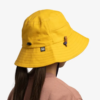 Una persona que lleva un Jockey Buff Niños 5 Panel Go Black Noru, un sombrero de pescador amarillo brillante con correa para la barbilla y etiqueta con el logotipo. La persona tiene cabello largo y oscuro y lleva una camisa malva sobre un fondo blanco.