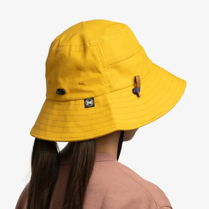 Una persona que lleva un Jockey Buff Niños 5 Panel Go Black Noru, un sombrero de pescador amarillo brillante con correa para la barbilla y etiqueta con el logotipo. La persona tiene cabello largo y oscuro y lleva una camisa malva sobre un fondo blanco.