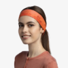 Una persona con cabello largo y castaño sonríe, vistiendo una vibrante diadema Cinta Buff CoolNet UV Slim Keffy Nectarine y una camiseta a juego, sobre un fondo blanco liso que resalta su actitud alegre.