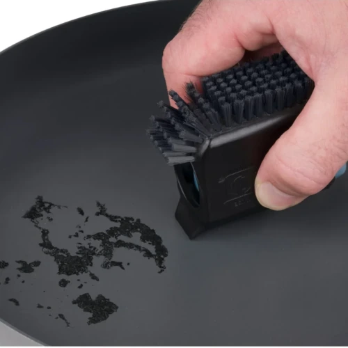 Una mano usa un cepillo rectangular con cerdas rígidas para frotar una mancha en una sartén del Set de Cocina Sea To Summit Detour 4pc (copia), revelando su superficie mayoritariamente lisa y de color gris oscuro.