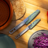 Dos elegantes cuchillos de cocina del Set de Cocina 4pc Sea To Summit Frontier (copia) con mangos verdes descansan sobre una superficie de madera con una estera tejida, una maceta de terracota y repollo morado en rodajas en un plato gris bajo una iluminación cálida.