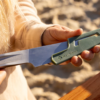 Una persona sostiene un cuchillo con una funda verde con la inscripción "Sea to Summit", probablemente del Set de Cocina de 4 piezas Sea To Summit Frontier (copia), sobre un fondo de arena y madera. Lleva una camisa beige de manga larga, que evoca un espíritu aventurero propio de la Frontera.