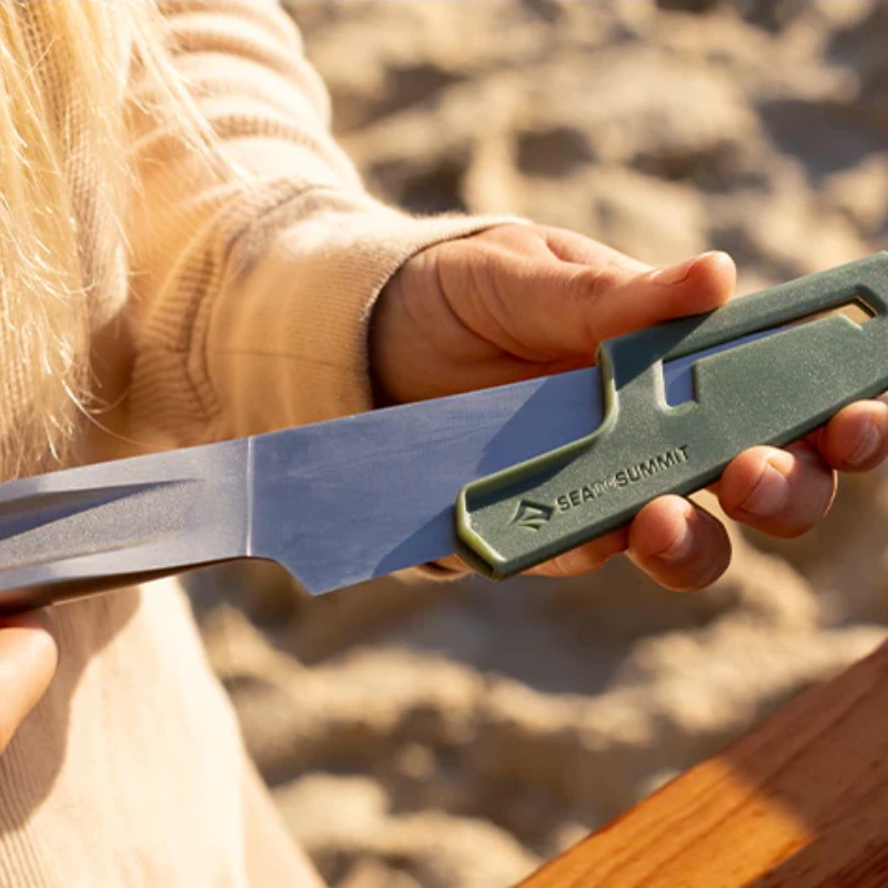 Una persona sostiene un cuchillo con una funda verde con la inscripción "Sea to Summit", probablemente del Set de Cocina de 4 piezas Sea To Summit Frontier (copia), sobre un fondo de arena y madera. Lleva una camisa beige de manga larga, que evoca un espíritu aventurero propio de la Frontera.