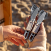 Una persona sostiene dos juegos de cubiertos metálicos plegables Sea To Summit, cada uno con una cuchara, un tenedor y un cuchillo de cocina Sea To Summit Detour (copia). La playa al fondo complementa su camiseta beige de manga larga para esta aventura costera.