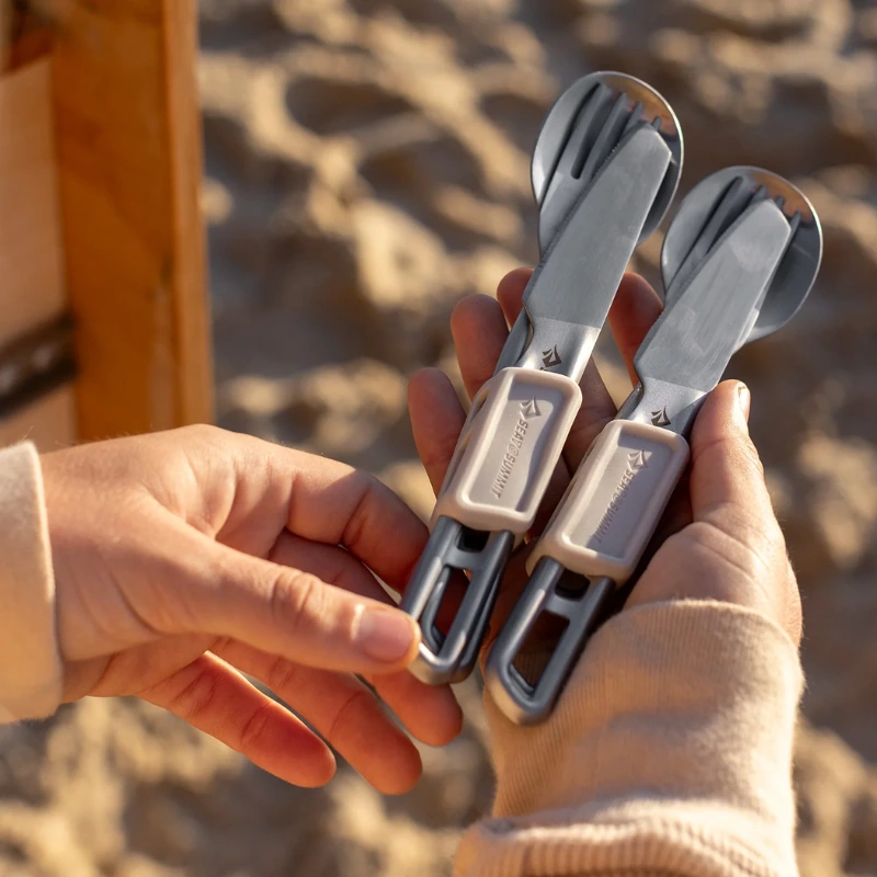Una persona sostiene dos juegos de cubiertos metálicos plegables Sea To Summit, cada uno con una cuchara, un tenedor y un cuchillo de cocina Sea To Summit Detour (copia). La playa al fondo complementa su camiseta beige de manga larga para esta aventura costera.