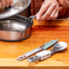 Una persona con chaqueta marrón levanta la tapa de una olla de acero inoxidable sobre una superficie de madera. Junto a ella, los utensilios de camping —un tenedor-cuchara y una navaja plegable con mangos verdes y blancos— están listos para la comida. Compañeros ideales para cualquier aventura de Sea To Summit Detour con Cuchillo de Cocina.