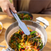 Utilizando el Set de Cocina Sea To Summit Detour 4pc, una persona usa pinzas para levantar fideos salteados con vegetales de una olla de metal repleta de ingredientes coloridos como pimientos morrones y verduras en una mesa de madera, mientras otro plato con contenido similar se encuentra en el fondo.