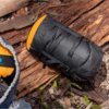Una mochila negra con detalles naranjas reposa en el suelo, entre hojas secas y ramas. A su izquierda, parte de un saco de dormir emerge de su elegante Bolsa de Compresión Sea To Summit Evac Heavy Duty Compression, en gris y naranja, integrándose a la perfección en esta escena al aire libre.
