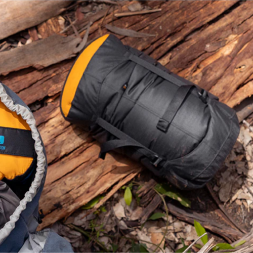 Una mochila negra con detalles naranjas reposa en el suelo, entre hojas secas y ramas. A su izquierda, parte de un saco de dormir emerge de su elegante Bolsa de Compresión Sea To Summit Evac Heavy Duty Compression, en gris y naranja, integrándose a la perfección en esta escena al aire libre.
