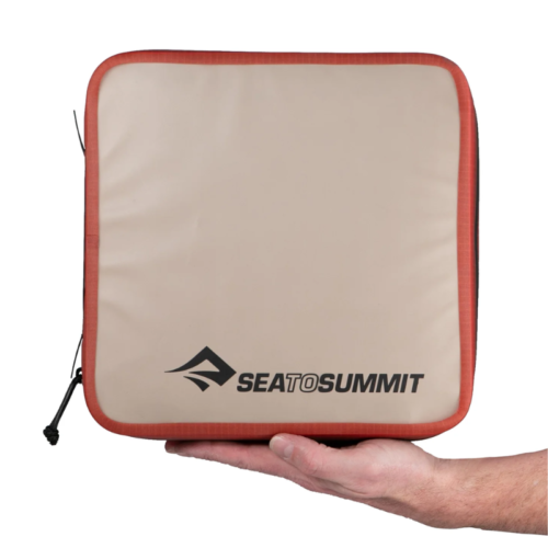 Una mano sostiene un estuche cuadrado beige y rojo con cremallera. El logo "Sea to Summit" se muestra audazmente en negro en la parte frontal, lo que sugiere su diseño robusto. Este cubo de embalaje hidráulico es perfecto para tus aventuras más exigentes.