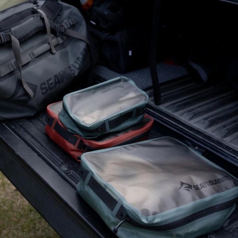 Cubos de embalaje Sea to Summit de varios tamaños y una bolsa de lona se organizan cuidadosamente en un vehículo. El Bolso Organizador Sea To Summit Hydraulic Packing Cube se presenta entre los artículos en verde, rojo y gris, mostrando un conjunto de equipo para actividades al aire libre.