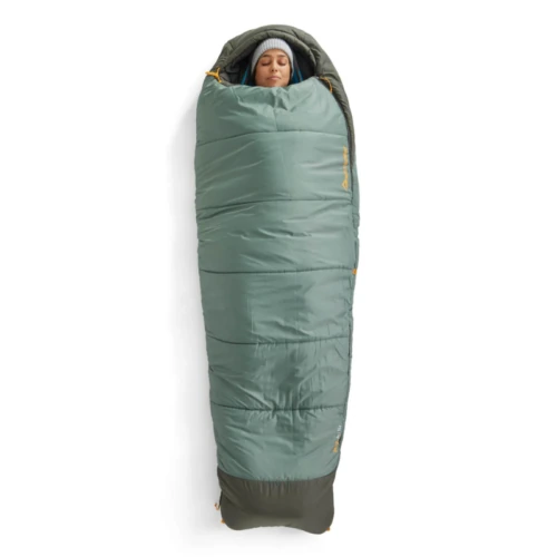 Una persona se acurruca al aire libre en un saco de dormir verde estilo momia, que evoca la calidez acogedora del Saco de Dormir Pluma Sea to Summit Ascent para mujer. Lleva un gorro, y la vibrante cremallera naranja añade un toque de color al fondo blanco.