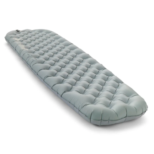 Se muestra una colchoneta inflable rectangular azul claro con un patrón acolchado sobre un fondo blanco. Vista desde un ángulo, la colchoneta está ligeramente elevada, con una válvula visible en la esquina superior.