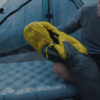 Una persona con camisa gris sentada sobre una colchoneta inflable Sea to Summit, ajustando un par de botines aislantes amarillos y grises dentro de una tienda de campaña. La tienda tiene una tela azul translúcida que permite la entrada de luz natural.