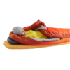 Una persona con un saco de dormir naranja está sobre una colchoneta Sea to Summit Pursuit Plus, con una camiseta amarilla, mangas grises y un gorro. Su cabeza reposa sobre una almohada gris con una parte del saco enrollada para mayor amortiguación.