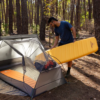 Una persona con camisa azul y gafas de sol está montando una tienda de campaña en el bosque. Dentro, coloca una colchoneta autoinflable Sea to Summit Pursuit Plus junto a una colchoneta naranja. Altos árboles los rodean, creando un sereno remanso al aire libre.