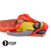 Una persona yace cómodamente en un saco de dormir rojo, con una camiseta amarilla y gris y un gorro, sobre una colchoneta autoinflable Sea to Summit Pursuit Plus naranja (copia). Un logo en la esquina dice "1% for the Planet - Member".
