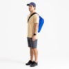 Una persona con camiseta beige, pantalones cortos grises y zapatillas negras está de pie de lado con una Correa Desmontable Sea To Summit para Bolsa Seca colgada del hombro. Lleva una gorra azul marino sobre un fondo blanco liso.