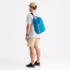 Una persona con una gorra beige, una camiseta gris, pantalones cortos beige y zapatillas negras está de lado con una Correa Desmontable Sea To Summit para Bolsa Seca azul con escritura amarilla colgada sobre un hombro sobre un fondo blanco liso.