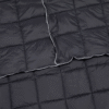 Un primer plano de la Manta térmica Outdoor Puelo Kano en gris oscuro, con acolchado visible, ribetes blancos contrastantes a lo largo de los bordes y un material texturizado acogedor, parcialmente doblado para mostrar su diseño.