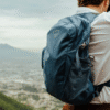 Una persona con una camisa blanca y una Mochila Osprey Tempest 20 Mujer (copia) azul está al aire libre con un paisaje urbano borroso y montañas en el fondo.