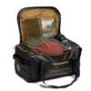 Una bolsa de viaje negra y marrón está abierta, revelando ropa, un par de zapatos y un libro titulado "Excursiones". La bolsa tiene asas negras y la palabra "ATAKAMA" impresa en dorado en un lateral.