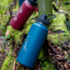 Botellas de agua de colores brillantes, entre las que destaca la elegante Botella Térmica Atakama Outdoor 1L en rojo, verde y azul, están dispuestas sobre corteza de árbol cubierta de musgo en un exuberante entorno forestal.