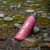 La botella térmica Atakama Outdoor de 710 ml en color rojo reposa sobre piedras mojadas junto a un arroyo, rodeada de guijarros suaves y agua que fluye, creando un ambiente sereno similar a un oasis.