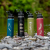 Cuatro Botellas Térmicas de 710 ml de Atakama Outdoor, con un diseño impactante, descansan sobre rocas húmedas junto a un arroyo. Las vibrantes botellas de azul, negro, verde y rojo, con tapas negras, están rodeadas de exuberante vegetación y agua que fluye suavemente al fondo.