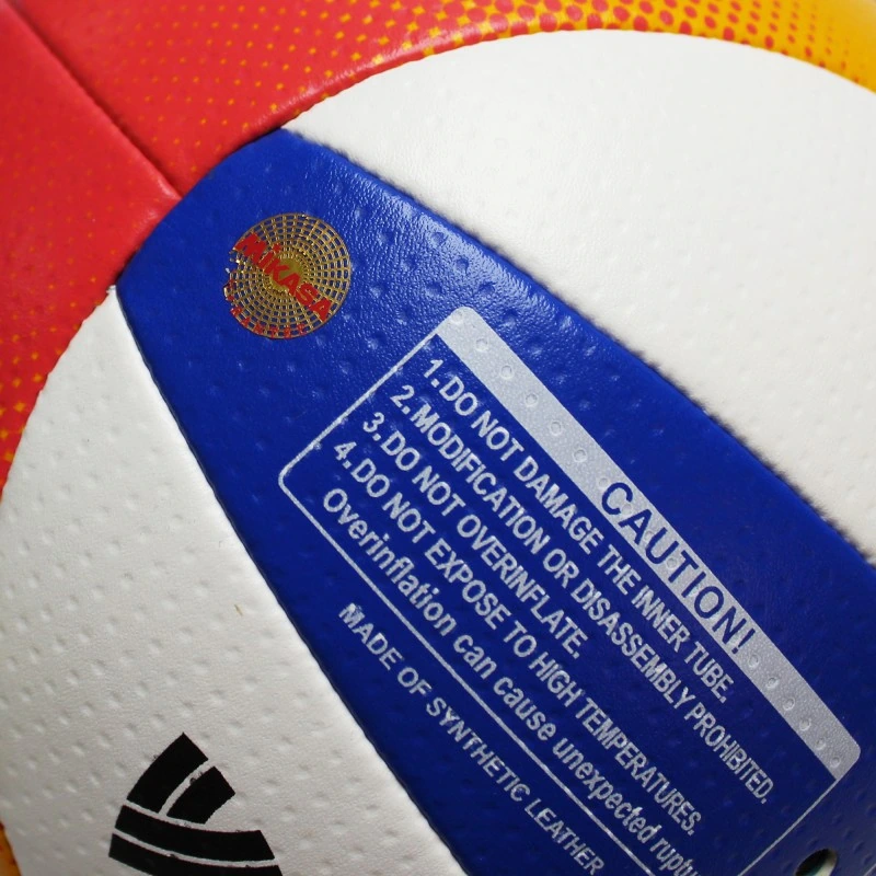 Primer plano de un Balón de Voleibol de Playa MIKASA BV550C con una etiqueta de advertencia que recomienda no dañar la cámara ni exponerlo a altas temperaturas. El balón es rojo, blanco, azul y amarillo con cuero sintético texturizado.
