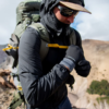 Un excursionista, con chaqueta con capucha y gafas de sol, se encuentra en un sendero rocoso de montaña. Lleva guantes térmicos Atakama Outdoor Isoflex Malleco y una mochila grande con correas amarillas. Lleva una gorra debajo de la capucha mientras el agreste paisaje de Atakama se extiende bajo el cielo nublado.