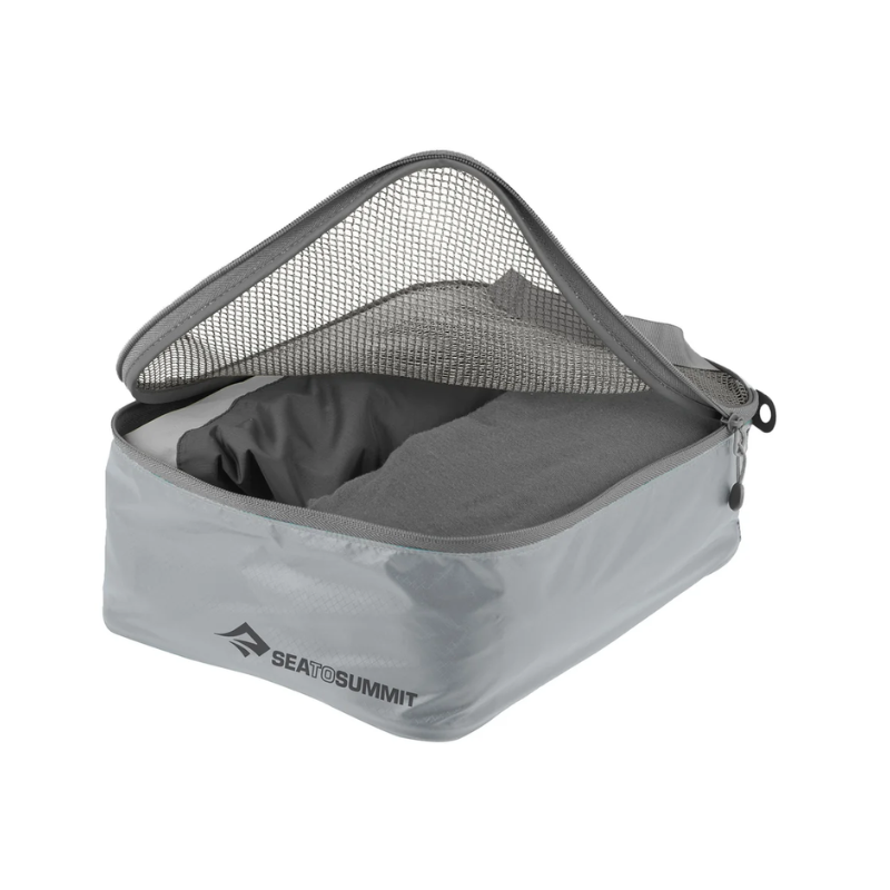 La Bolsa Organizadora Sea To Summit Garment Mesh es un cubo de embalaje gris con costuras redondeadas y una tapa de malla, que se abre parcialmente para revelar ropa perfectamente doblada. La inscripción "Sea to Summit" se muestra de forma prominente en el lateral, lo que la hace perfecta para viajes organizados.